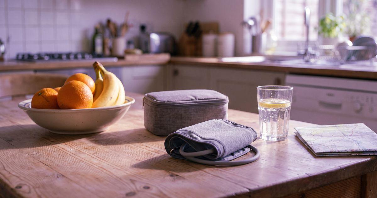 A coiled blood pressure cuff by its case, a bowl of oranges and bananas, sparkling water and a folded map on a sunny table.