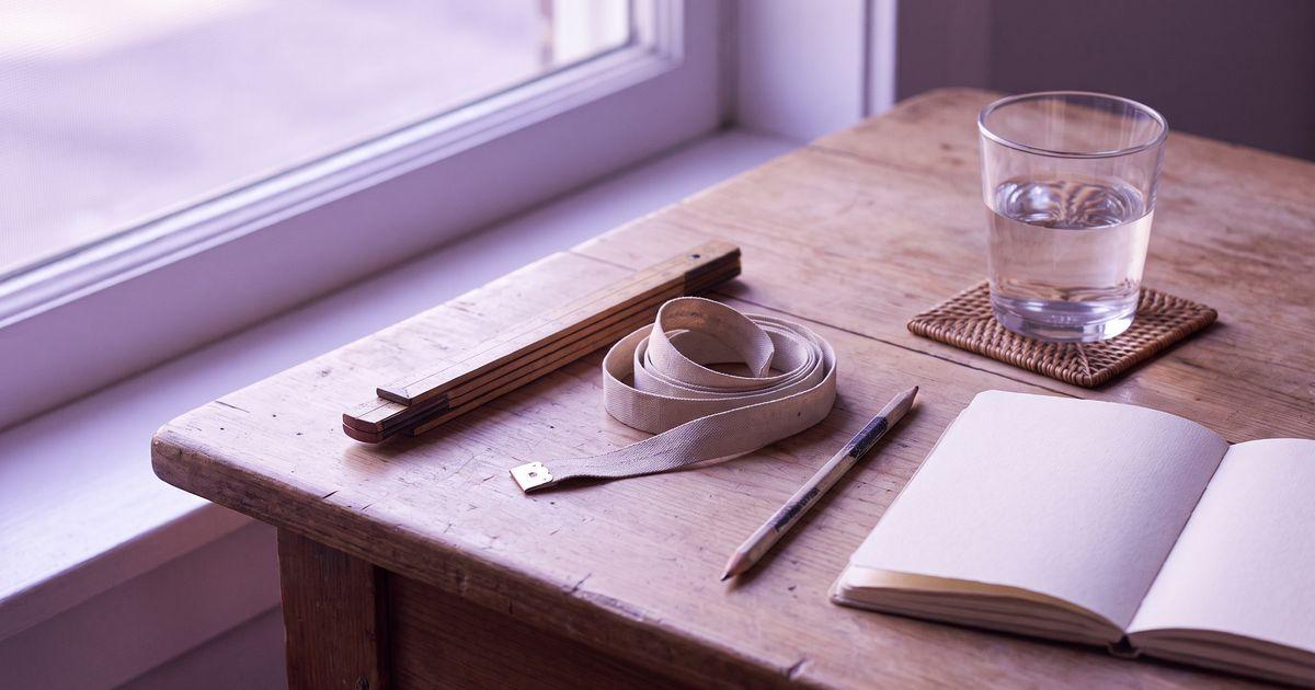 A wooden folding ruler, a coiled cloth tape, a pencil, a notebook and a glass of water on a sunlit desk.