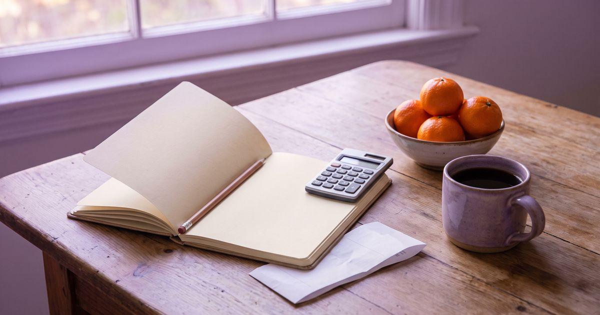An open budget notebook with a pencil, a calculator, a mug of coffee and a bowl of clementines on a sunlit kitchen table.