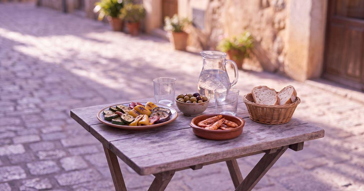 A café terrace table with grilled vegetables, olives, grilled prawns, a small bread basket and a water jug in late afternoon light.