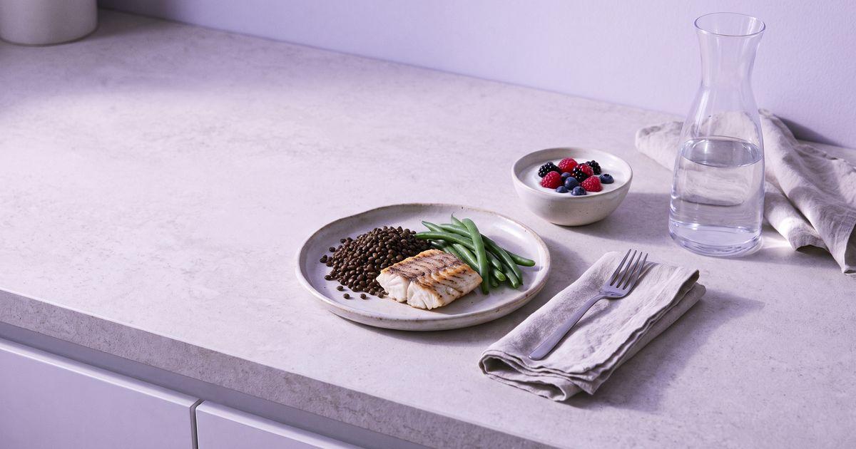 A small plate of grilled fish, lentils and green beans with a bowl of yoghurt and berries and a water carafe on a stone counter.