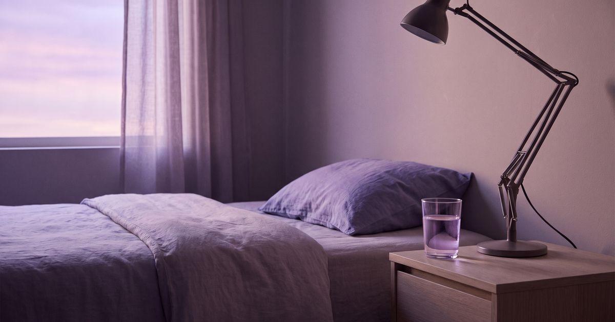 A made bed with a crumpled pillow, a glass of water and a lamp on the bedside table, pale dawn light at the window.