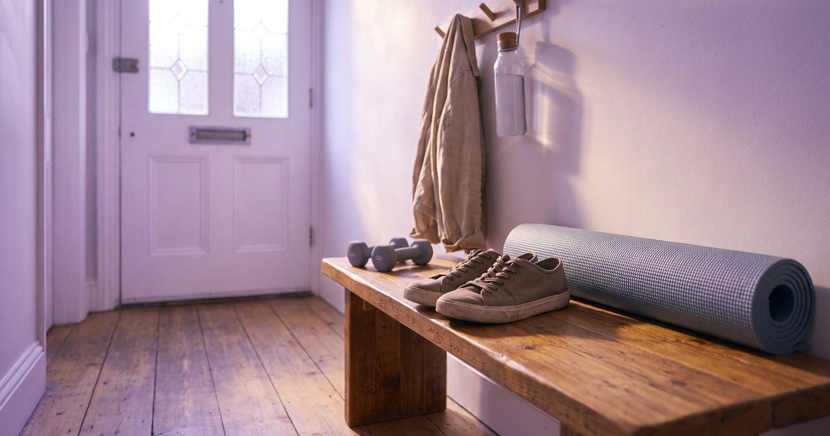 Chaussures de marche, tapis de sport roulé et deux petits haltères posés sur un banc en bois près d'une porte d'entrée, avec une veste accrochée au portemanteau.